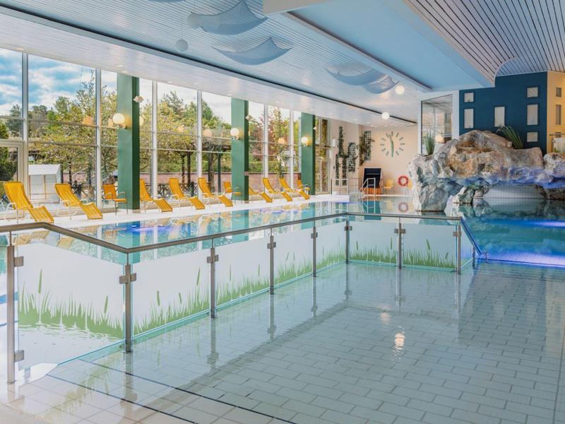 Helles Hallenbad mit Glasfassade, Liegestühlen und dekorativem Felsenbereich.