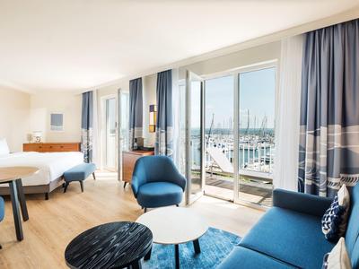 Modern hotel room with balcony overlooking marina, blue-white decor, light wooden floors.