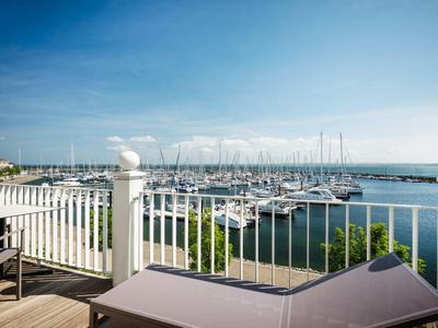 View of a marina with sailboats and lounge chairs on a terrace by the water.
