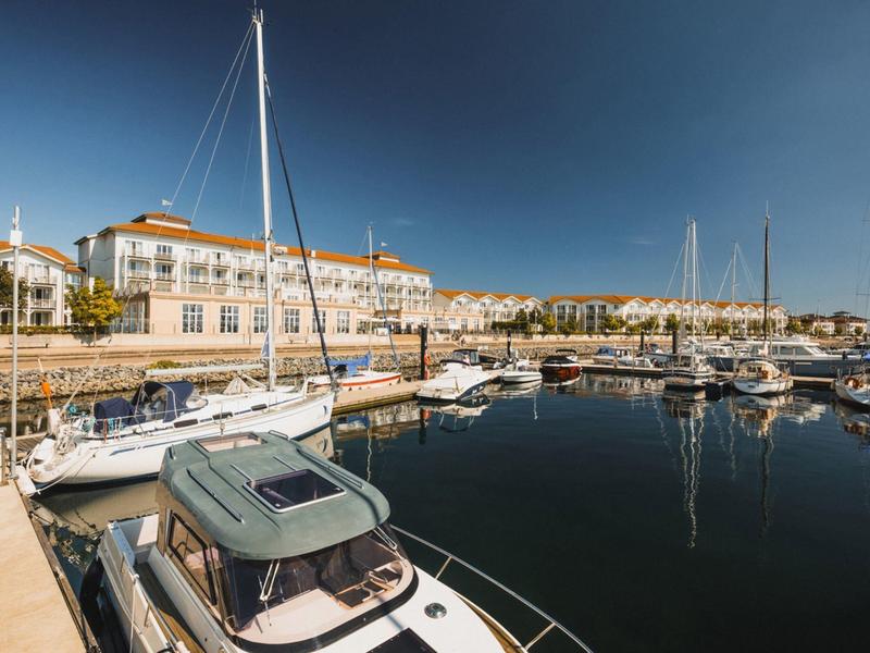 Sailboats and yachts docked in a calm marina in front of a hotel under a clear sky.