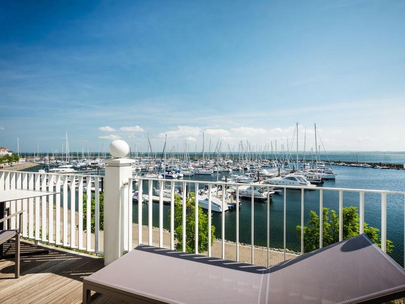 View of a marina with sailboats and lounge chairs on a terrace by the water.