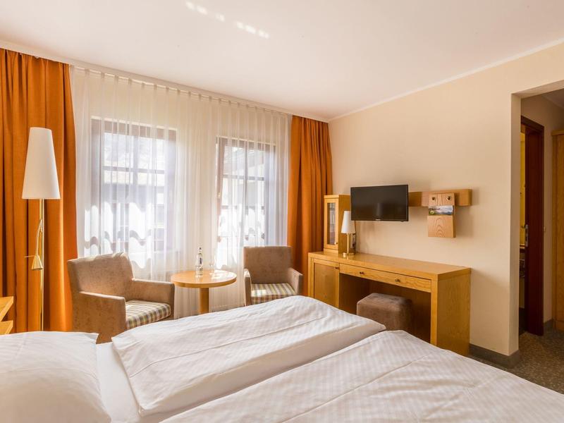 Chambre d'hôtel lumineuse avec deux lits, rideaux orange et coin salon près de la fenêtre.