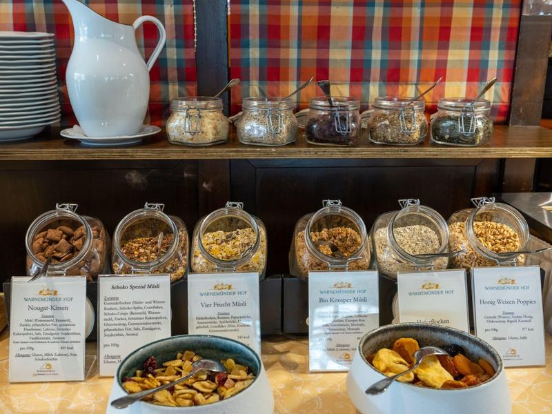 Différents céréales de petit-déjeuner et fruits secs dans des bocaux avec descriptions sur un buffet.