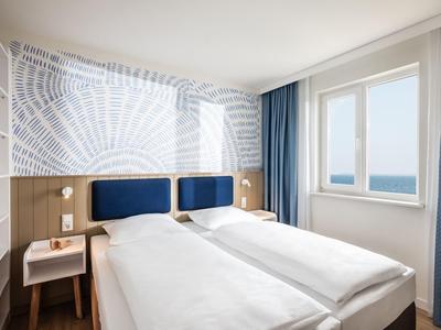 Chambre d'hôtel lumineuse avec deux lits simples, têtes de lit bleues et fenêtre avec vue sur mer