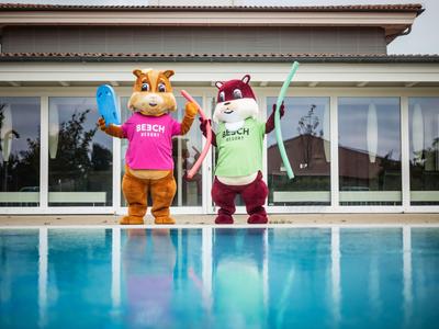 Deux mascottes colorées se tiennent près d'une piscine devant un bâtiment avec de grandes fenêtres.