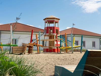 Aire de jeux avec sol sableux et structure d'escalade devant un bâtiment aux toits rouges.