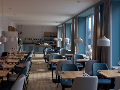 Moderní restaurace s modrými křesly, dřevěnými stoly, velkými okny a uvolněnou atmosférou.