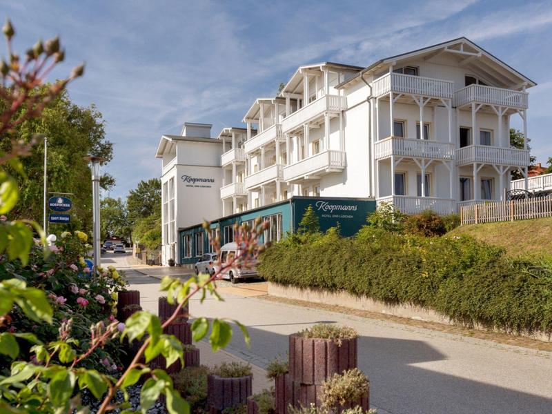 Modernes weißes Hotelgebäude mit Balkonen und Pflanzen am Straßenrand unter blauem Himmel.