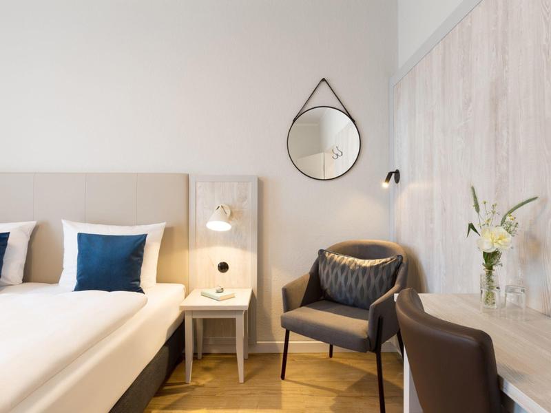 Modernes Hotelzimmer mit zwei Betten, Nachttisch, Sessel und großem Spiegel an hellen Wänden.