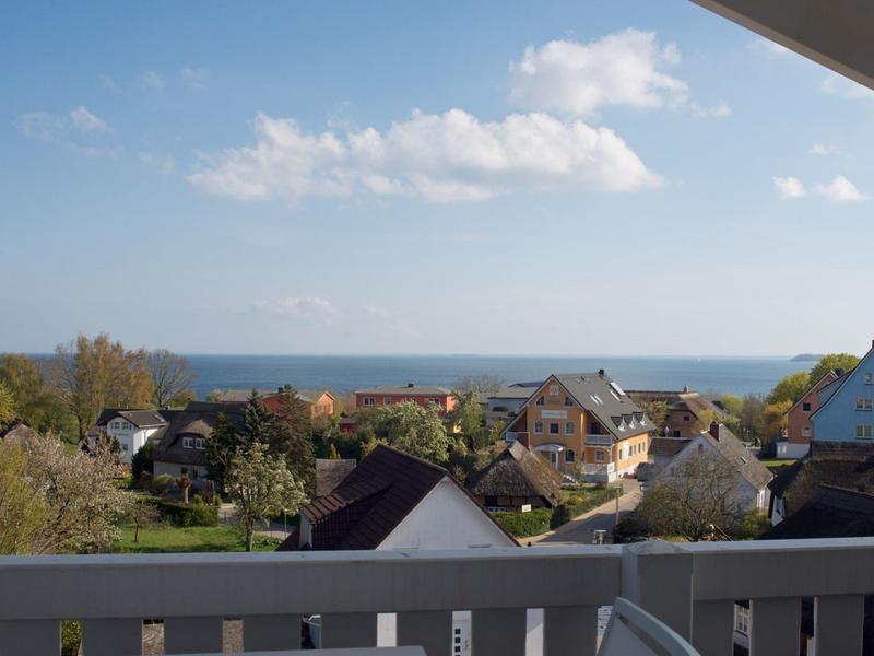 Blick von einem Balkon auf ein Dorf mit Häusern und dahinter das Meer unter blauem Himmel.