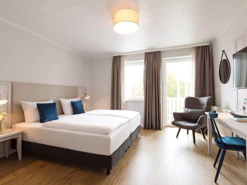 Helles Hotelzimmer mit Doppelbett, Sesseln, Schreibtisch und Balkon mit Blick ins Grüne.