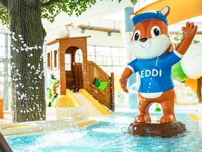 Kleurrijk kinderspeelgebied met glijbaan en mascottefiguur in levendig binnenzwembad.