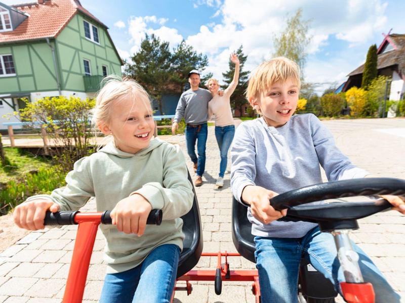 Twee kinderen rijden lachend op een tweepersoonsvoertuig in een zonnig vakantiepark.