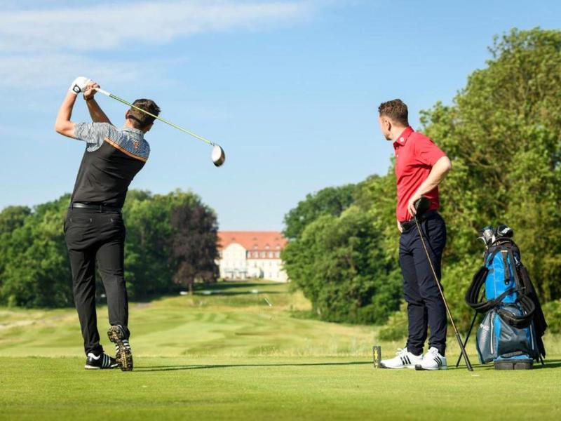 Twee mannen op een golfbaan, een slaat de bal terwijl de ander toekijkt.