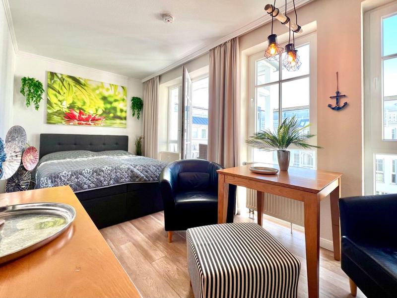 Světly hotelový pokoj s manželskou postelí, dřevěnou podlahou, psacím stolem, křesly a velkými okny.