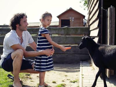 Uomo e bambina che danno da mangiare a una capra davanti a una fattoria con un capannone di legno