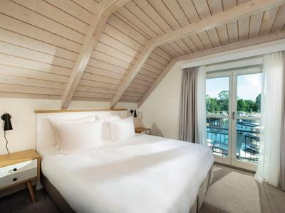 Gemütliches Schlafzimmer mit weißer Bettwäsche, Holzbalkendecke und Blick auf Balkon und grüne Umgebung.