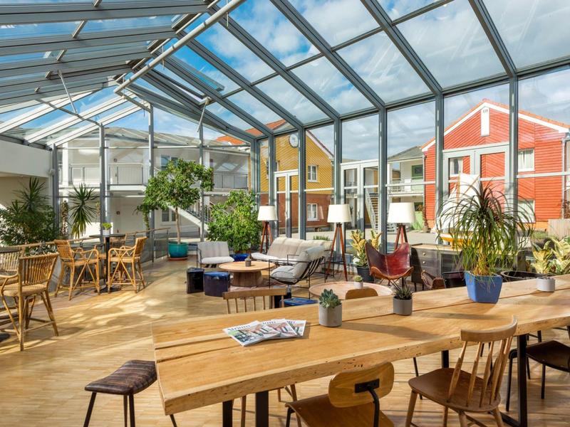 Bright conservatory with wooden and rattan furniture, plants, and colorful houses in the background.