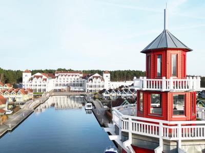 Blick auf einen Hafen mit einem roten Leuchtturm und weißen Gebäuden im Hintergrund unter blauem Himmel.