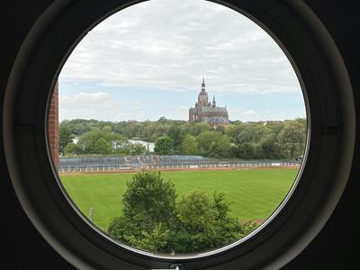 Widok przez okrągłe okno na zielone pole i historyczny zamek w tle.