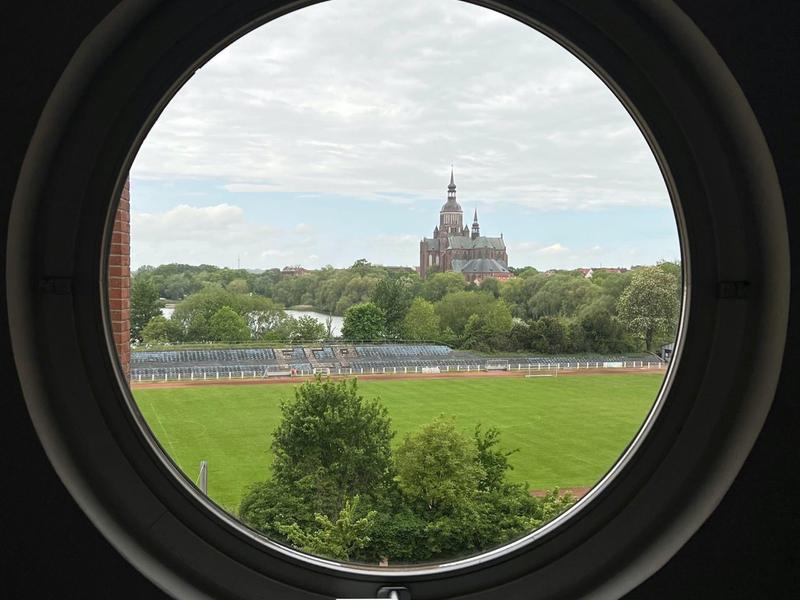 Widok przez okrągłe okno na zielone pole i zamek w tle.