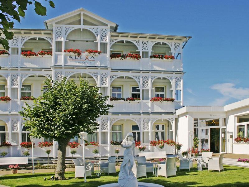 White three-story building with balconies and flowers, below a garden with trees and statue.
