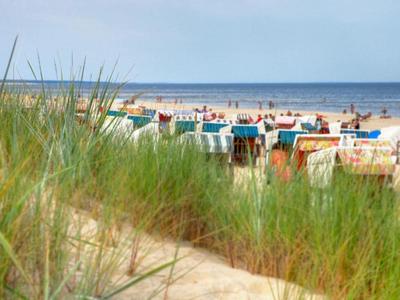 Pohled přes písečné trávy na pláž s barevnými plážovými křesly a lidmi u moře.