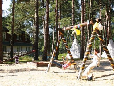 Děti si hrají na houpačce v písku s lesem a indiánskými stany v pozadí.