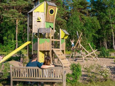 Pár sedí na lavičce před prolézačkou a skluzavkou v lesním hřišti.