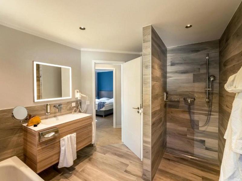 Moderní koupelna s dřevěným vzhledem, bezbariérovým sprchovým koutem, umyvadlem a otevřenými dveřmi do ložnice.