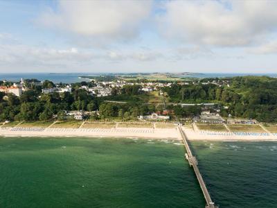 Vue aérienne d'une longue jetée s'étendant jusqu'à une plage de sable dans une ville côtière boisée.