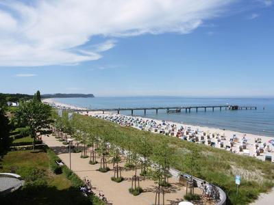 Strandpromenade mit Bäumen, Strandkörben und Holzsteg am Meer unter blauem Himmel.