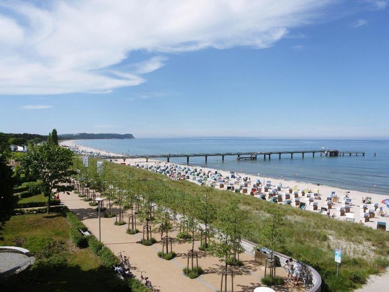 Strandpromenade mit Bäumen, Strandkörben und Holzsteg am Meer unter blauem Himmel.