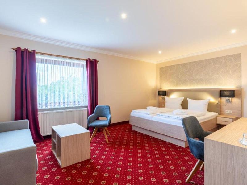 Bright hotel room with double bed, armchair, desk, and red carpet.