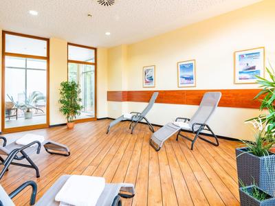 Heller Ruheraum mit Liegestühlen, Holzboden und großen Fenstern zu einem Balkon mit Blick auf Palmen.