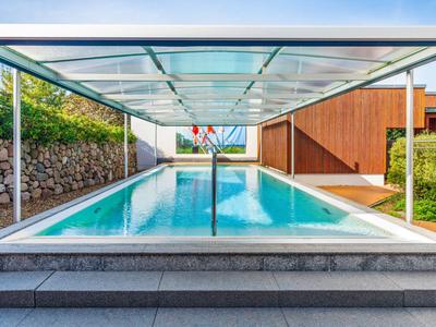 Moderner Pool mit Glasüberdachung und Basketballkorb neben Holzgebäuden und Garten.