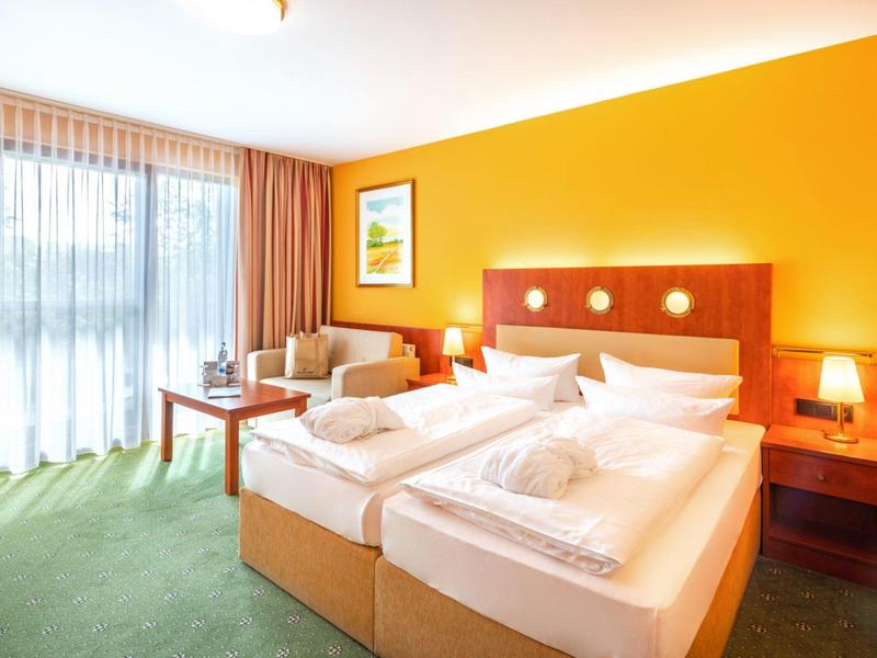 Hell eingerichtetes Hotelzimmer mit Doppelbett, Sofa, großem Fenster und orangefarbener Wand.