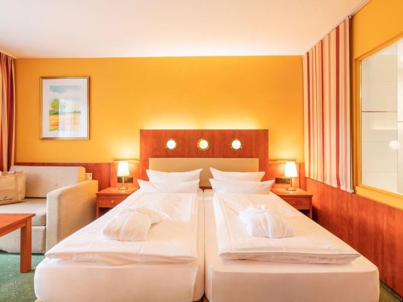 Helles Hotelzimmer mit zwei Einzelbetten, orangener Wand und einem Sitzbereich mit Sofa.