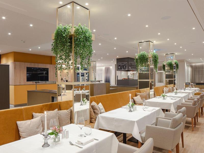 Modern restaurant with white tables, comfortable chairs, and hanging plants in a hotel.