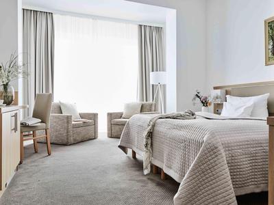 Jasny pokój hotelowy z dwoma łóżkami pojedynczymi, dwoma fotelami i dużym oknem z zasłonami.