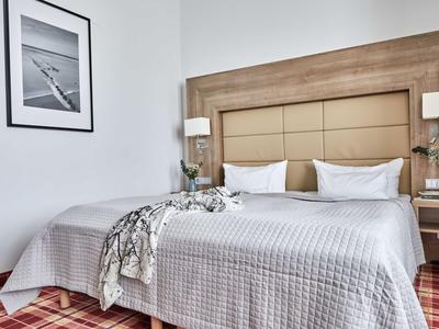 Nowoczesny pokój hotelowy z łóżkiem podwójnym, drewnianym zagłówkiem i dekoracyjnym obrazem na ścianie.