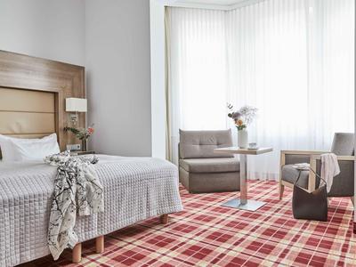 Jasny pokój hotelowy z łóżkiem podwójnym, fotelami, małym stolikiem i kratowaną wykładziną.