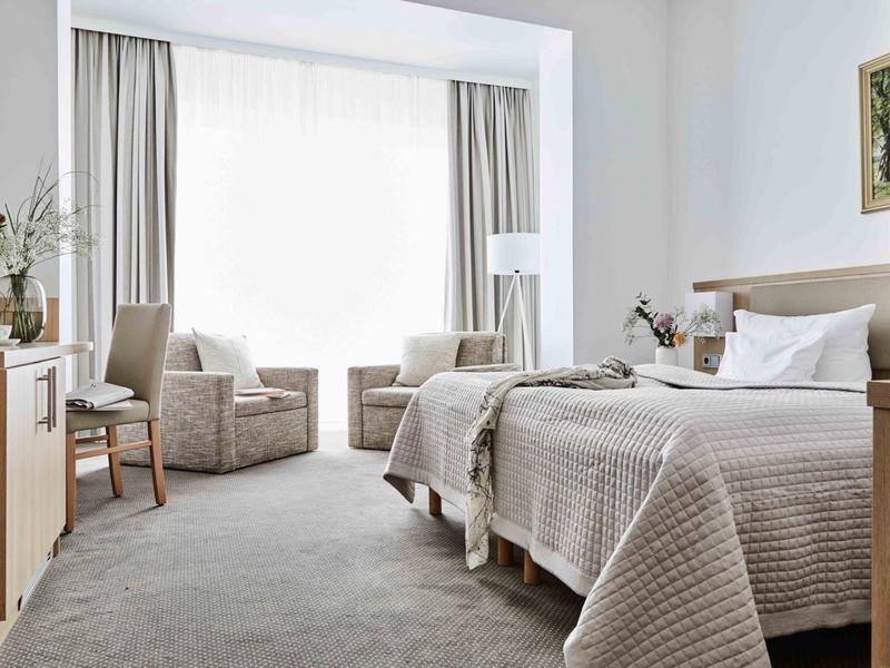 Jasny pokój hotelowy z dwoma pojedynczymi łóżkami, fotelami i oknem z zasłonami.