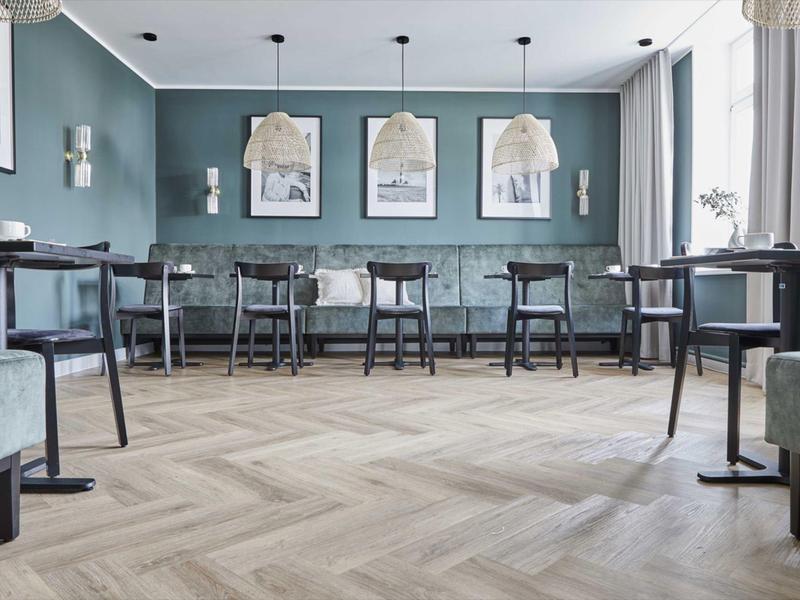 Nowoczesna strefa wypoczynkowa w hotelu z niebieskimi ścianami, drewnianymi krzesłami i jasną podłogą w jodełkę.