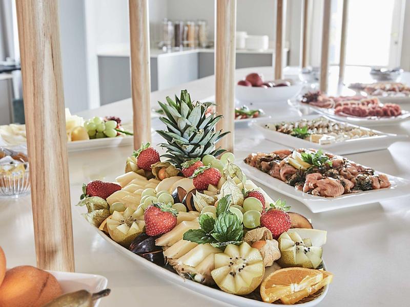 Bufet z różnymi owocami i daniami śródziemnomorskimi na stołach w jasnym pomieszczeniu.