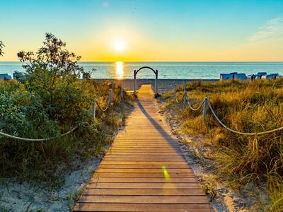 Sentiero di legno attraversa le dune verso la spiaggia con tramonto sul mare.