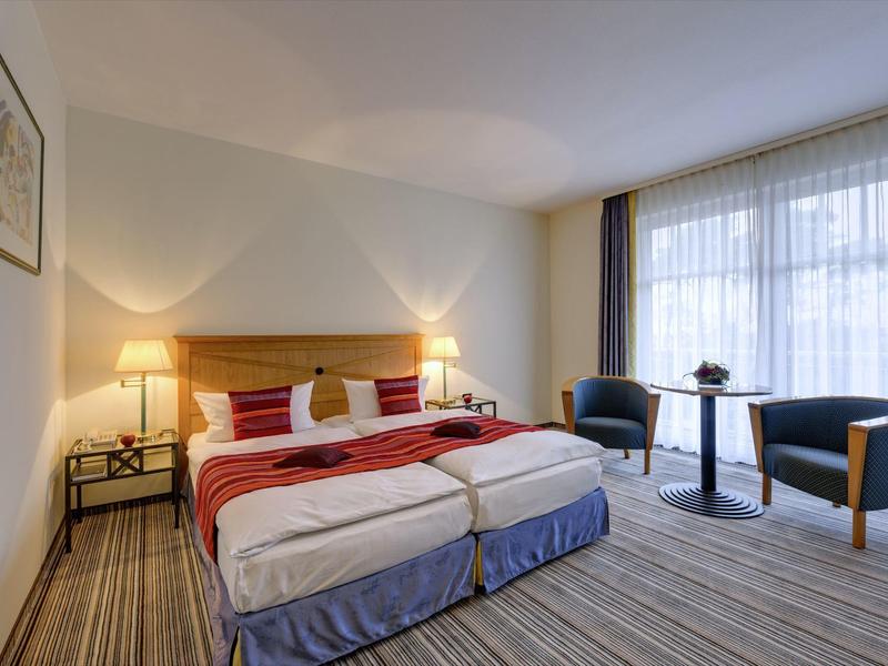 Hotel room with two single beds, bedside lamps, and seating area by large windows.