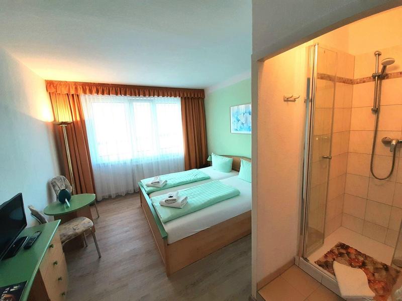 Hell eingerichtetes Schlafzimmer mit Doppelbett, Dusche und TV, in warmen Farbtönen gestaltet.