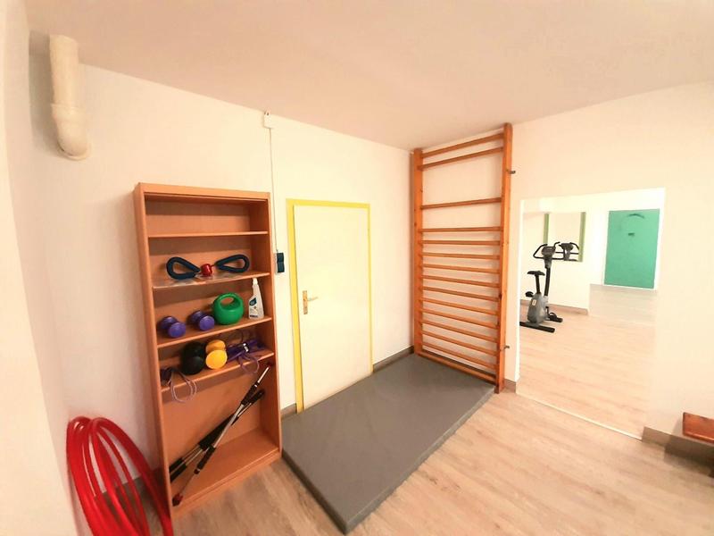 Ein heller Fitnessraum mit Regal, Gymnastikmatte, Sprossenwand und Trainingsgeräten im Hintergrund.