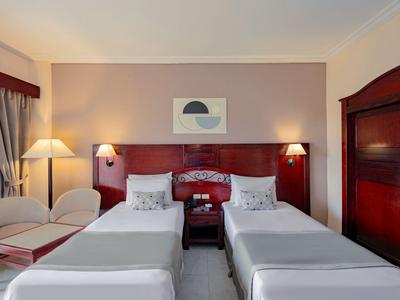 Hotelzimmer mit zwei Einzelbetten, Nachttisch, Stühlen und weicher Beleuchtung.